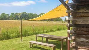 Un banc et une table en bois sont installés sous un auvent jaune, dans un champ herbeux, avec une clôture en arrière-plan.