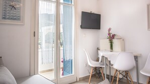 Una camera moderna con una porta scorrevole in vetro che dà su un balcone, una TV a schermo piatto montata a parete, un tavolo bianco con un vaso di fiori e sedie bianche.