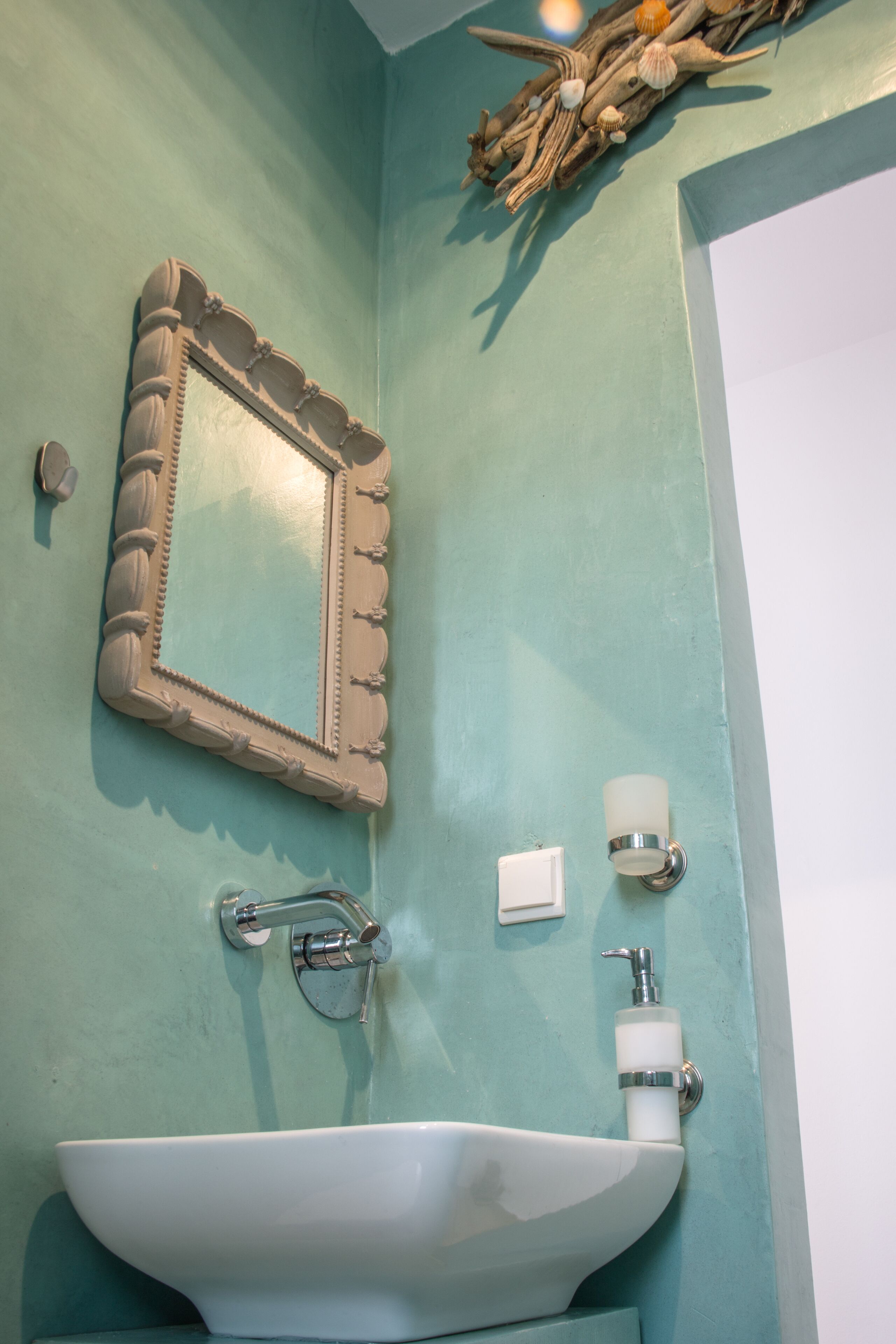 Un baño con pared verde azulado, un lavamanos blanco, un espejo decorativo y un dispensador de jabón en la pared.