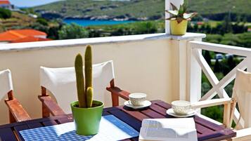 A wooden table with a book, a cup, and a cactus in a green pot on a balcony overlooking a coastal landscape.