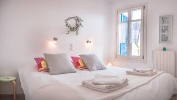 A neatly made bed with white linens and a few colorful pillows, a small bedside table with a lamp, and a window with blue shutters.