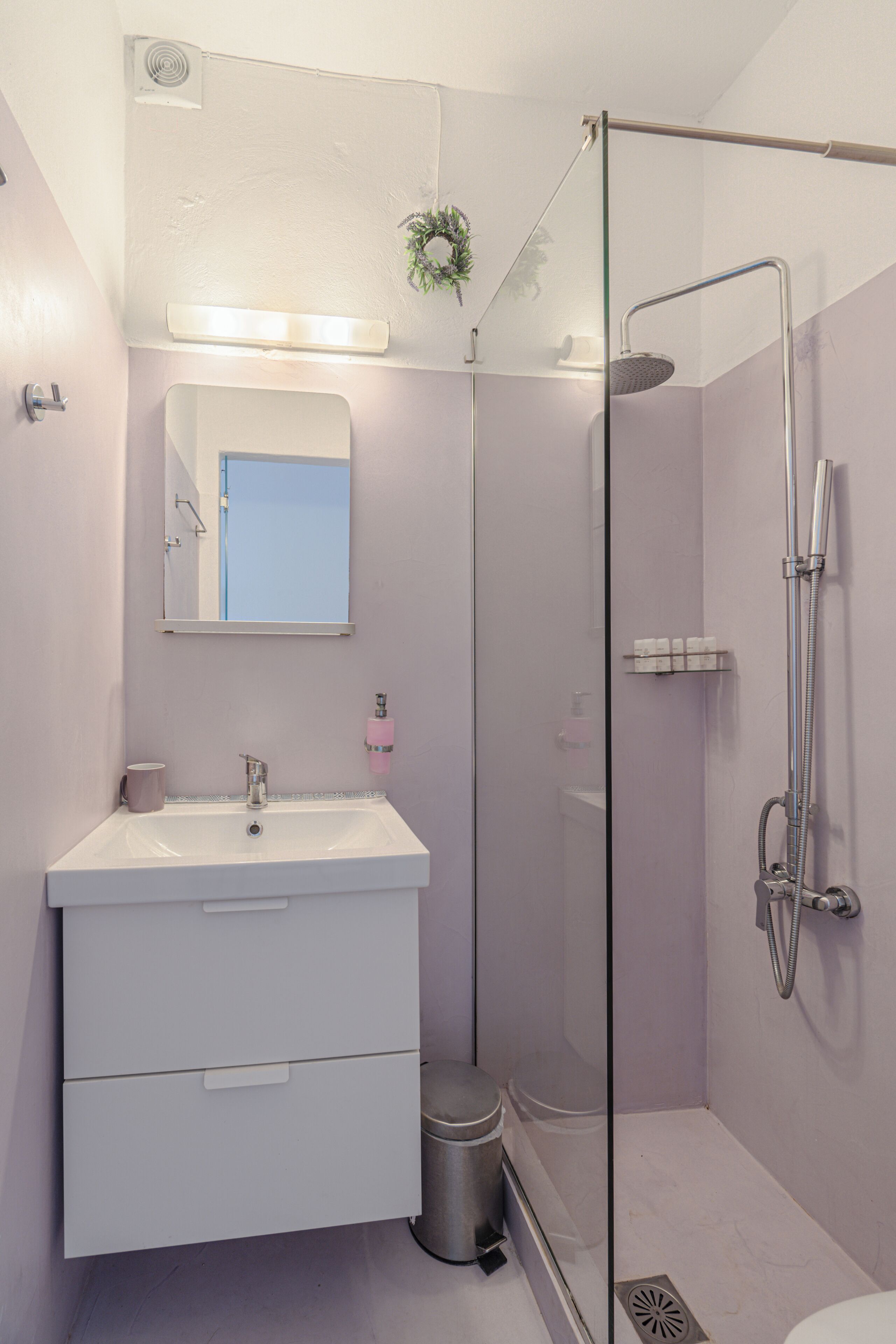 Un baño con ducha envuelta en vidrio, un lavamanos con espejo y un cubo de basura.