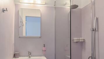 Un baño con ducha envuelta en vidrio, un lavamanos con espejo y un cubo de basura.