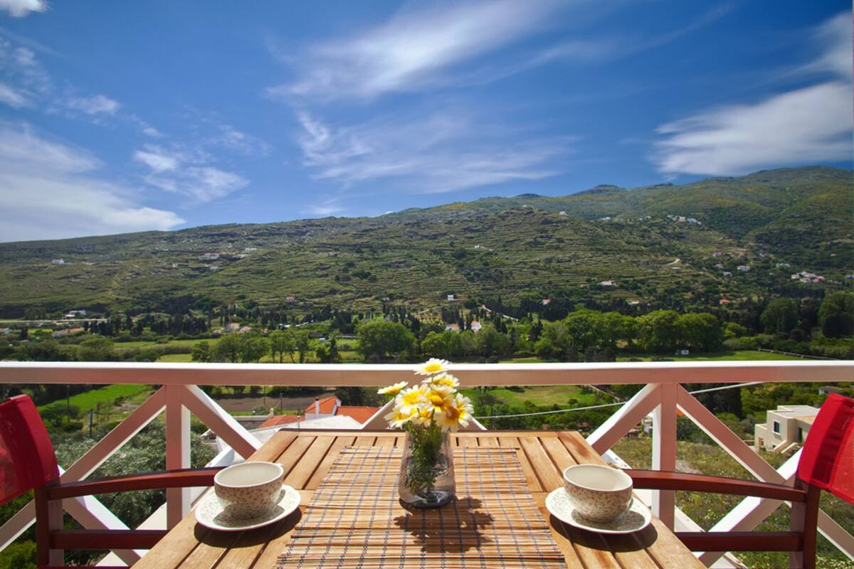 Una mesa de madera con un florero, dos tazas y vistas a colinas verdes y casas.