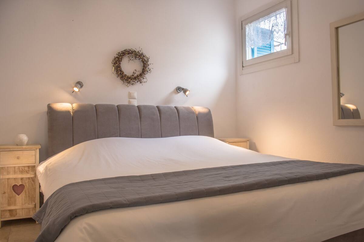 Un dormitorio con cama, mesitas de noche, una lámpara de pared, una ventana y un espejo.
