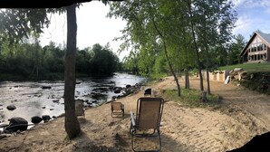 A lakeside cabin with a deck, surrounded by trees and a sandy beach.