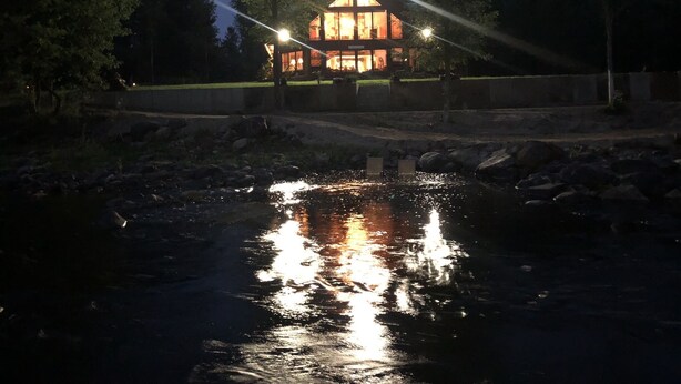 Uma cabana com fachada de vidro, rodeada por árvores, refletindo a luz na água.