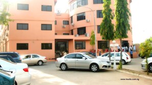 A multi-story building with a peach-colored facade, several windows, and a sign indicating it is a hotel. There are cars parked in front and a few people near the entrance. - K Suite Hotels (Kano)