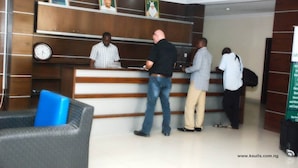 A reception area with a wooden reception desk, a clock, and a blue chair. - K Suite Hotels (Kano)