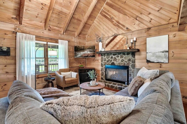A cozy log cabin interior with a stone fireplace, wooden walls, a comfortable sofa, and a television.