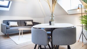 Un salon moderne avec un canapé gris, une table ronde accompagnée de deux chaises, une petite table basse blanche et un grand vase contenant des plantes séchées.