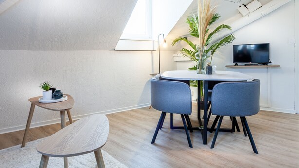 Une pièce moderne et bien éclairée, avec une table à manger, deux chaises, une petite table d’appoint ornée d’une plante et un téléviseur posé sur une étagère.