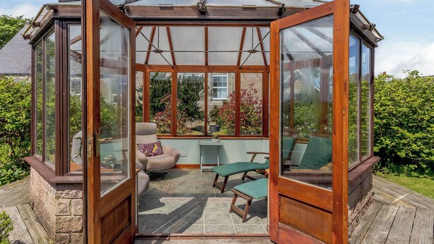 Une véranda dotée d’une structure en bois, de grandes surfaces vitrées et d’un sol carrelé. À l’intérieur, on trouve deux fauteuils verts rembourrés et une table en bois.