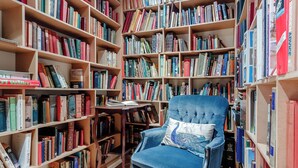 Une bibliothèque avec des étagères remplies de livres, un fauteuil bleu avec un coussin et une table en bois sur laquelle repose un livre.