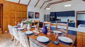 Un coin repas avec une table en bois dressée pour six personnes, une cuisine avec des meubles bleus et des poutres en bois au plafond.