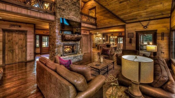 A rustic cabin interior with a stone fireplace, leather sofas, a dining area, and a kitchen. - The Haven on Toccoa - River deck | Fire Pit | Hot Tub | Outdoor Fireplace (Blue Ridge)