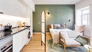 A compact apartment with a kitchenette, a bed with a green wall, and a window providing natural light. - Boavista Green Studio by DA'HOME (Porto)