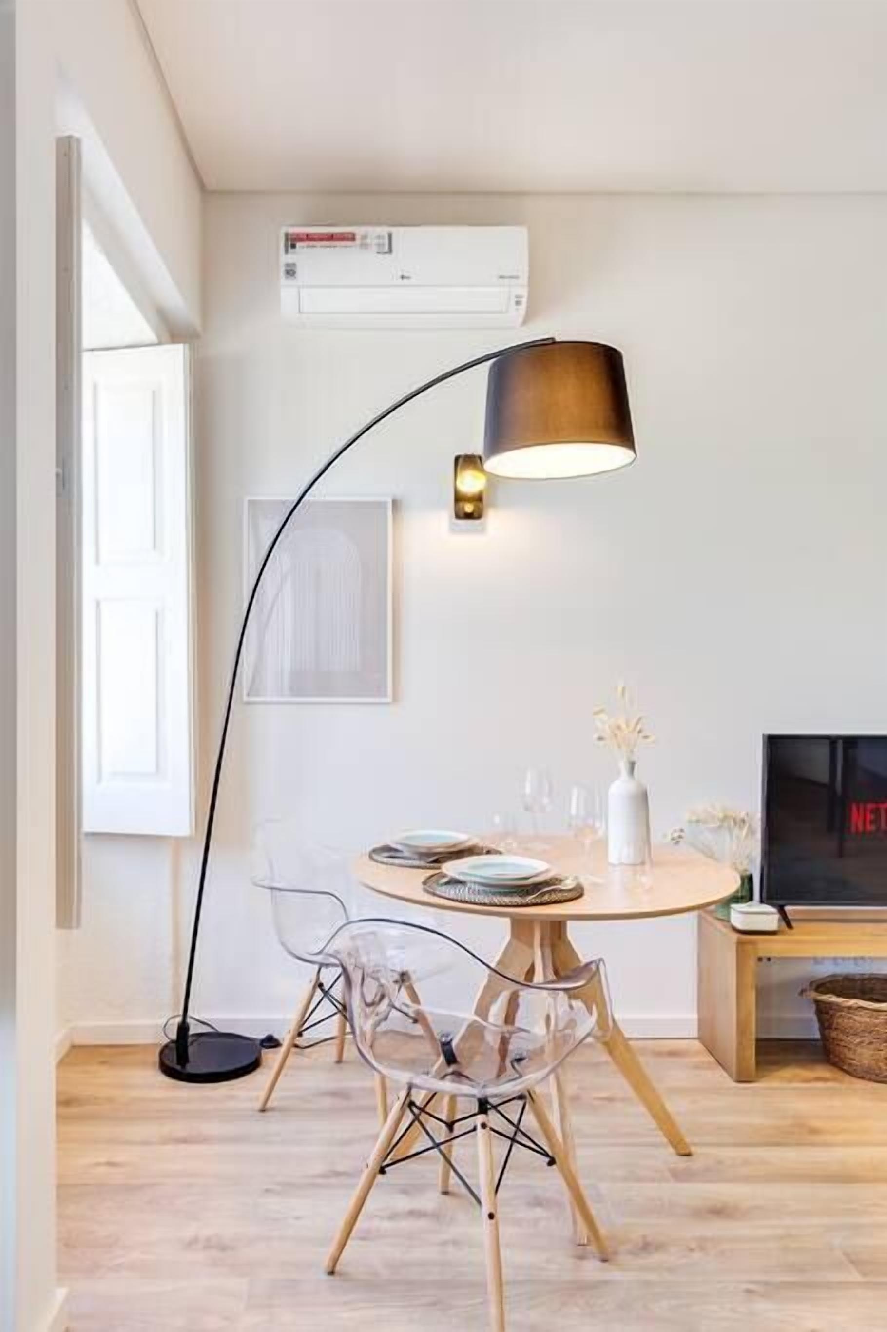 A dining area with a round table, clear chairs, a black floor lamp, a white vase with flowers, and a flat-screen TV displaying 'NETFLIX'.