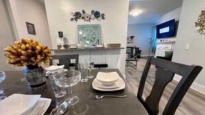 Een eetruimte met een donker houten tafel gedekt voor vier personen, inclusief borden, glazen en bestek. Op de tafel staat een vaas met gele bloemen, en erboven bevindt zich een decoratief object aan de muur.