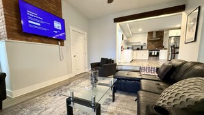 A modern living room with a flat-screen TV, leather sofa, glass coffee table, and a view into the kitchen. - Large 1BR on hill close to everything in Pitt (Pittsburgh)