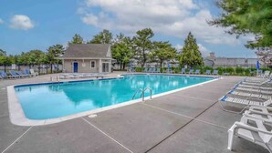 En rektangulär swimmingpool med ett klart, blått vatten, omgiven av en stenlagd terrass. Runt poolen finns solstolar placerade, och i bakgrunden syns en liten byggnad med en blå dörr.
