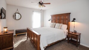 A bedroom with a wooden bed, a nightstand, a bedside lamp, a ceiling fan, a round mirror, and a window with curtains. - Patriots Point a Stylishly decorated duplex -Near MCAS Cherry Point– VERY CLEAN! (Havelock)