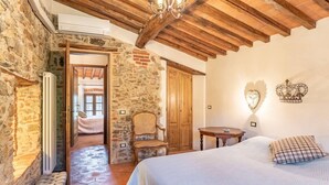 A bedroom with a stone wall, wooden ceiling beams, a bed with a plaid pillow, a chair, and a small table. - Villa Pera by Nicola Real Estate (Corsanico)
