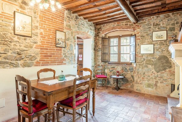 Une pièce rustique avec un mur en pierre, des poutres en bois au plafond, une table à manger avec des chaises et une petite table ronde surmontée d’un coussin rouge.
