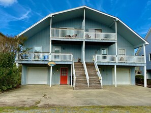 A two-story building with a front entrance, a red door, and a staircase leading to the second floor. - Coastal Distancing | Recently Updated | Under 5-min Walk to the Waves (Holden Beach)