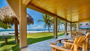 Un espace extérieur aménagé, abrité et doté de chaises et de tables en bois, offrant une vue sur une plage bordée de palmiers et sur l’océan.
