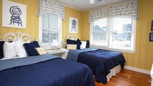 Habitación de hotel con dos camas, cada una con ropa de cama azul marino y cabeceras blancas. Hay dos ventanas con persianas, y las paredes están pintadas de amarillo.