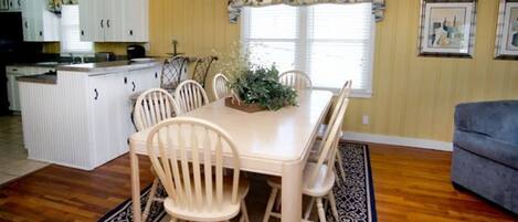 A dining area with a table and chairs, a kitchen in the background, and a living room with a couch.