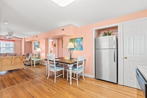 A kitchen with a wooden table, white chairs, a refrigerator, and a dining area with a sofa and a lamp.