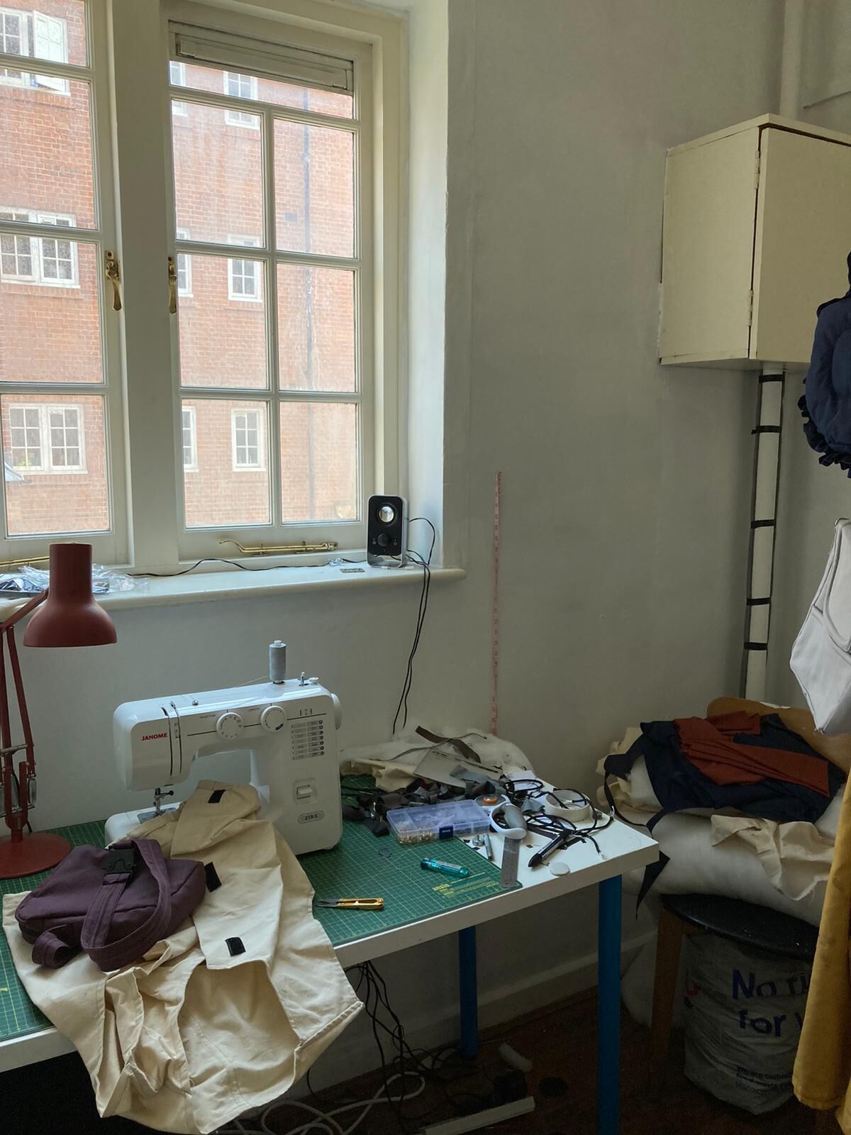 A sewing room with a sewing machine, a window with a view of brick buildings, a lamp, and various sewing materials on a table.