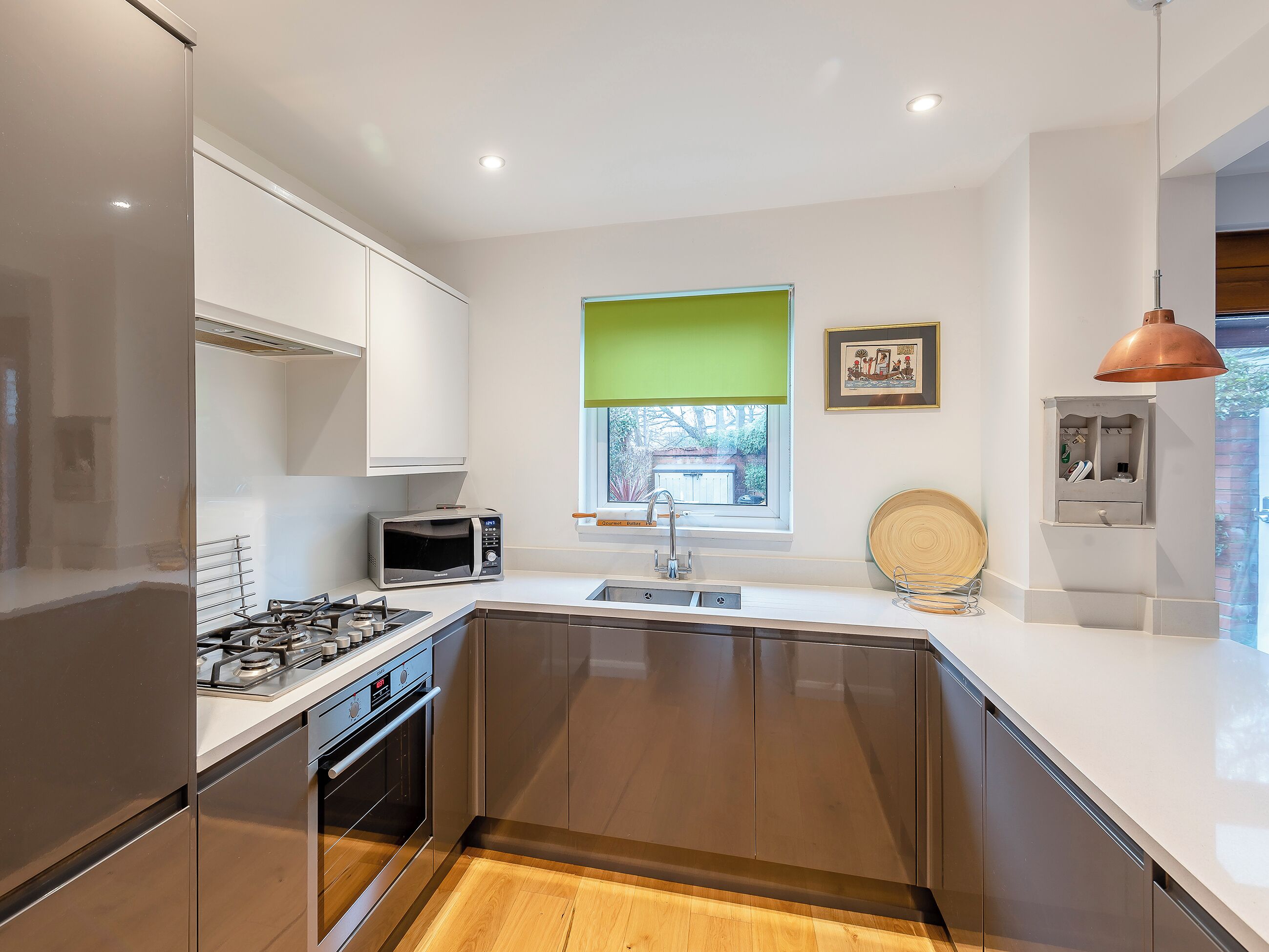 A modern kitchen with stainless steel appliances, a window with a green blind, and a wooden floor.