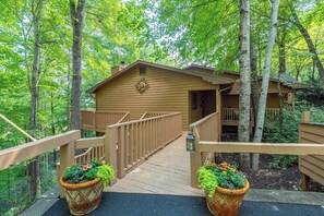 A wooden cabin with a wooden deck and a covered porch, surrounded by trees.