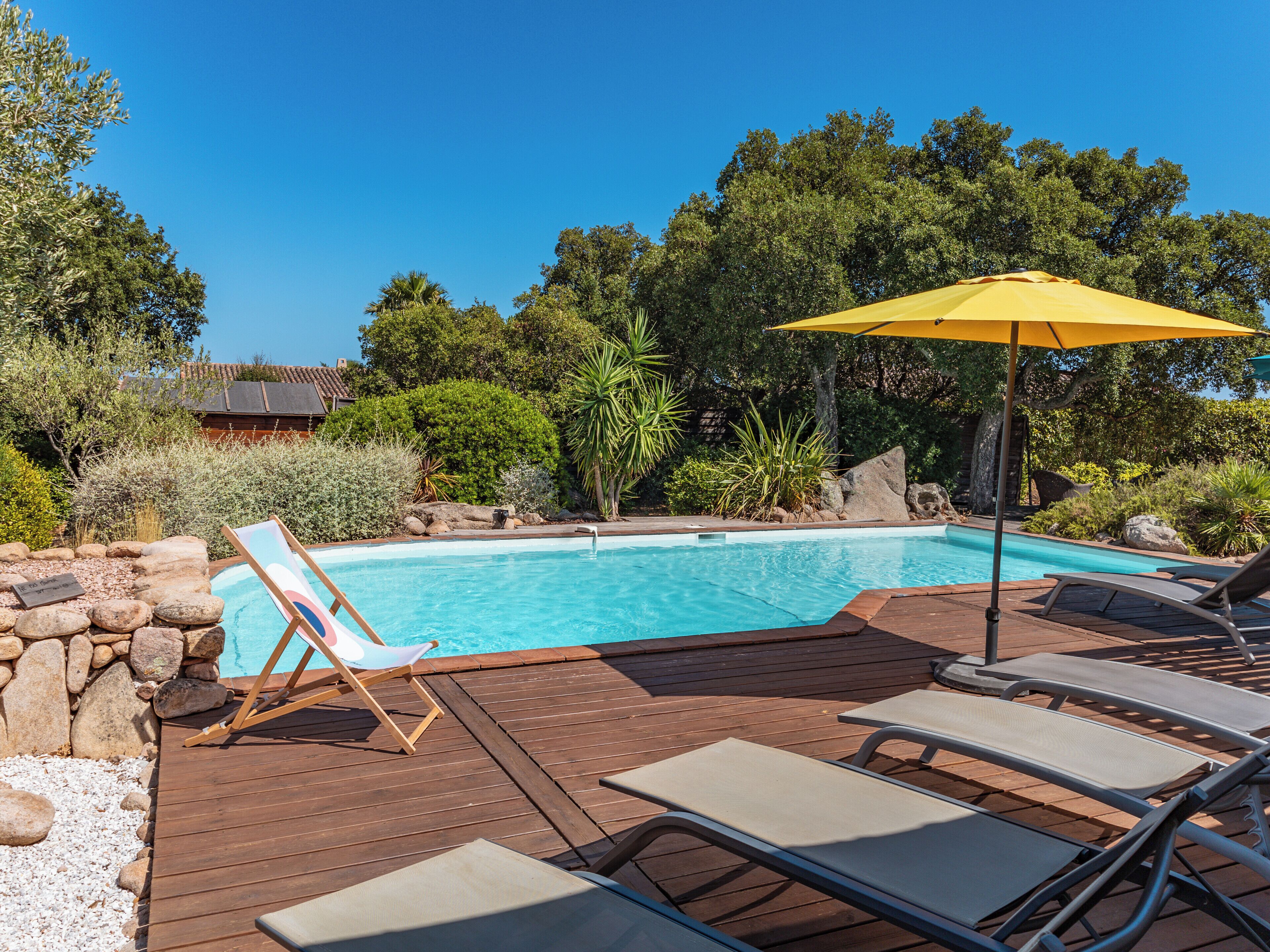 Um deck de madeira com piscina, cadeiras de descanso e um guarda-sol amarelo.
