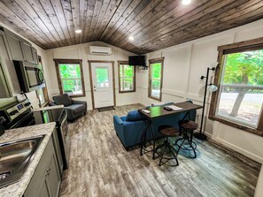 Una cocina moderna con horno y microondas integrados, una zona de comedor con mesa de madera y sillas azules, y ventanas amplias que ofrecen vistas a la vegetación.