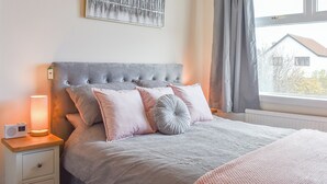 A bedroom with a tufted headboard, a bedside table with a lamp, and a window with curtains. - Och Moo at Number 47 (Seahouses)