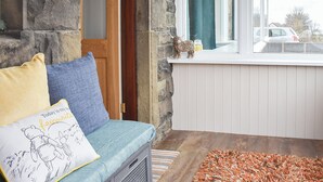 A cozy room with a stone wall, a wooden door, a window with a curtain, a blue sofa with cushions, and a rug on the floor. - Och Moo at Number 47 (Seahouses)