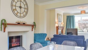 A living room with a fireplace, blue sofas, a dining table with chairs, and a large wall clock. - Och Moo at Number 47 (Seahouses)
