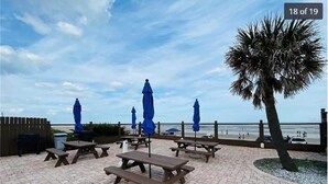 Eine Terrassenanlage im Freien mit Picknicktischen und blauen Sonnenschirmen mit Blick auf einen Strand.
