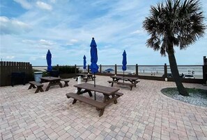 An outdoor patio with picnic tables and blue umbrellas, overlooking a beach. - Cozy Just Renovated Clean Ocean View Condo  Private Beach Access + Pools WiFi (Daytona Beach)