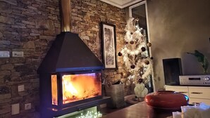 Een stenen muur, een open haard met vlammen, een lijst met een afbeelding, een kerstboom met lichtjes, en een televisietoestel op een meubel.