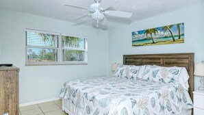 Un dormitorio con una cama, ventilador de techo, ventana con persianas, un cuadro de una playa y una mesita de noche con lámpara.