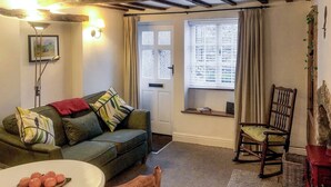 A cozy living room with a green sofa, a wooden rocking chair, a dining table with a plate of tomatoes, and a window with curtains. - 1 bedroom accommodation in Kirkby Lonsdale (Kirkby Lonsdale)