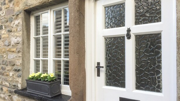 A stone building with a white door marked with the number 10, a window with shutters, and a planter with yellow flowers. - 1 bedroom accommodation in Kirkby Lonsdale (Kirkby Lonsdale)