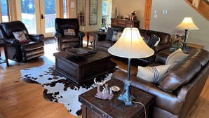 A spacious living room with a leather sofa, a leather armchair, a wooden coffee table, and a lamp. There is a cowhide rug on the floor and a staircase in the background. - Red River, NM Upper Valley Mountainside Retreat (Red River)