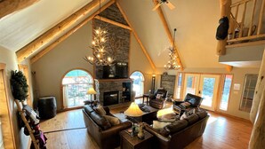 A spacious loft-style living room with a stone fireplace, wooden beams, and a large window offering a view of the outdoors. - Red River, NM Upper Valley Mountainside Retreat (Red River)