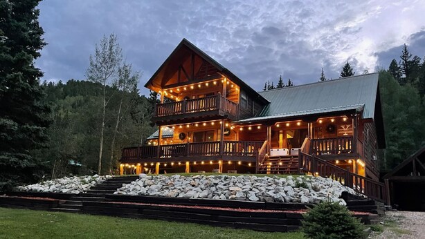 A two-story log cabin with a balcony and string lights, surrounded by a stone retaining wall and a gravel path. - Red River, NM Upper Valley Mountainside Retreat (Red River)